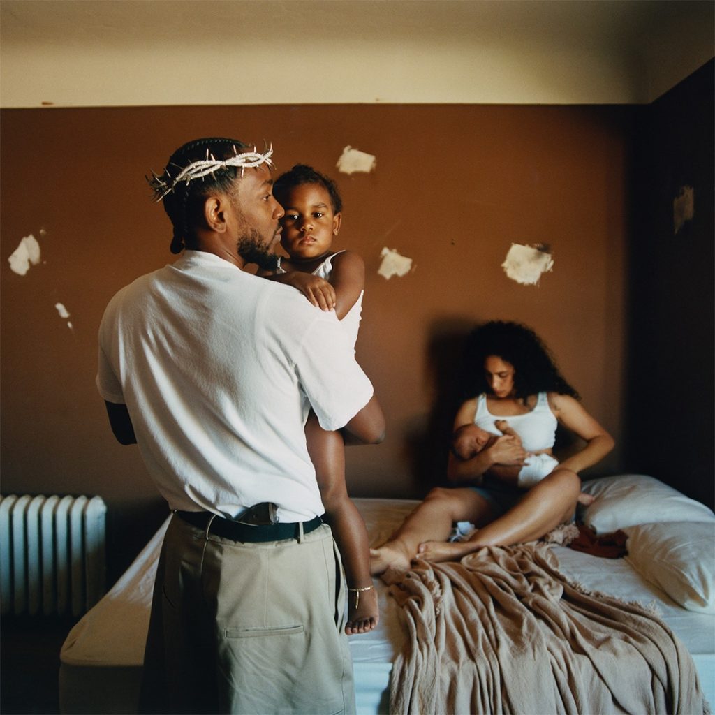 A black man in a crown of thorns and the handle of a gun in his waistband holds a complacent child in his arms while a woman breastfeeds on a bed.