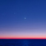 A dark sky with a few scattered stars fades to a bright pink horizon over dark waters