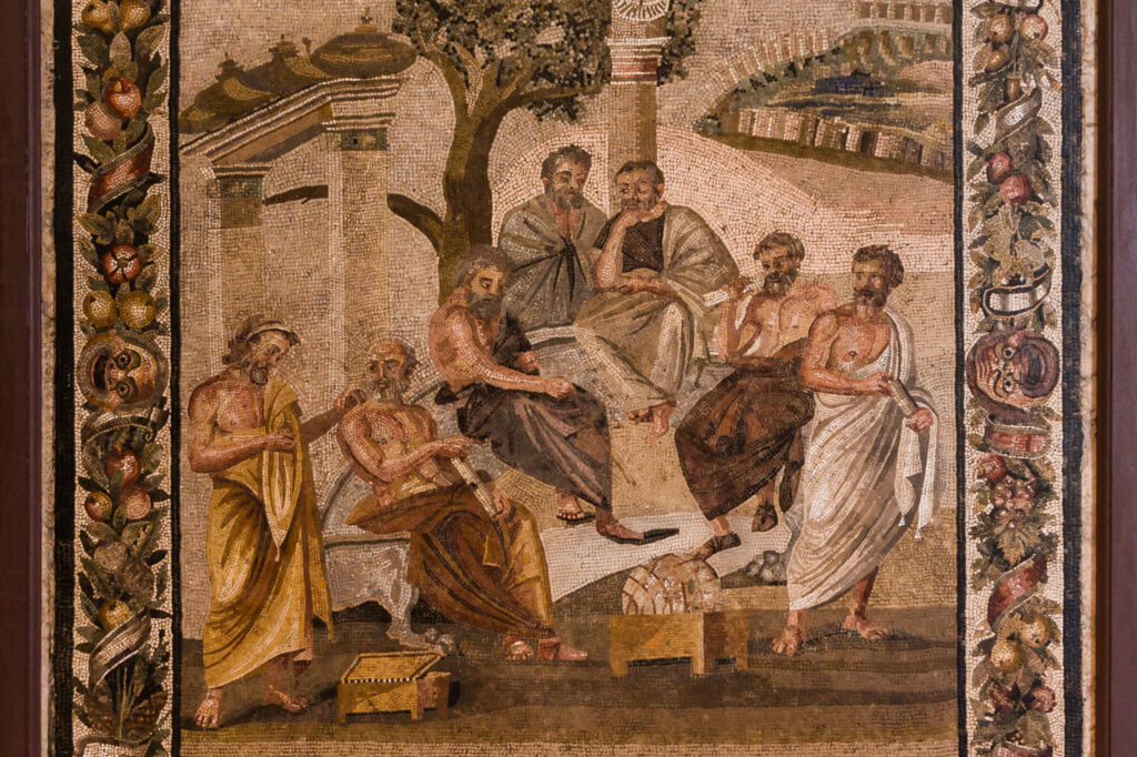 A mosaic of Socrates and six other men in himations lounging outside. Socrates is using a stick to point to the ground, and the men seem to be thinking and talking.