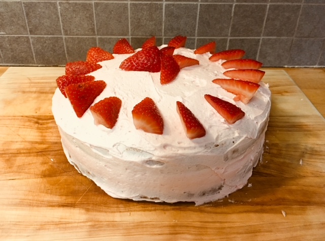 A photograph of a cake. The cake is placed on the middle of a wooden table. It is covered with light pink frosting and adorned with a circle of quartered strawberries. Imperfections make it clear that the cake is homemade.