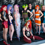 Eight people pose outdoors in front of a black tarp. They are dressed in a variety of bright and pastel colors, including brightly colored hair. Each outfit has a monochromatic color theme, and the outfits feature fishnet tights, sequins, and sea-inspired flourishes.