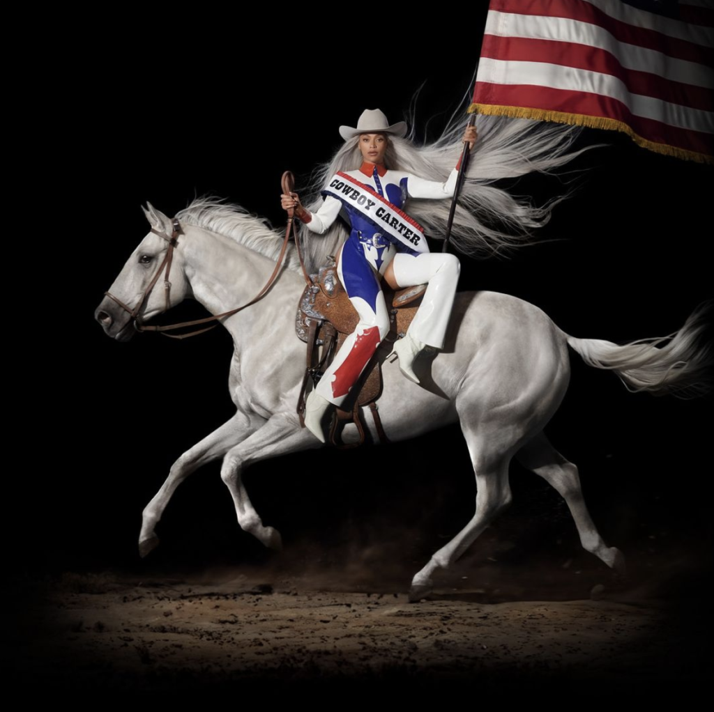 Beyoncé faces the camera in a shiny red, white, and blue cowboy-style suit and white cowboy hat. She rides side-saddle on a white horse in motion, her hair flowing out and matching the horse's mane. In one hand, she carries the reins, and in the other, a large American flag, of which only the bottom left quarter is visible. The background is black.