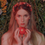 Natalie Wynn - head and shoulders - faces the camera directly, appearing nearly nude. She artfully holds an apple with the fingertips of both hands, nails red to match. Blond hair tumbles down out of a flower headband, all set against a green and floral background.