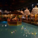 A dark room designed to look like it is outdoors. It has a large pool of fenced-in water in the center with a fake boat containing a live band. Bamboo accents, dried grass "roofs", plants, and string lights decorate the space.